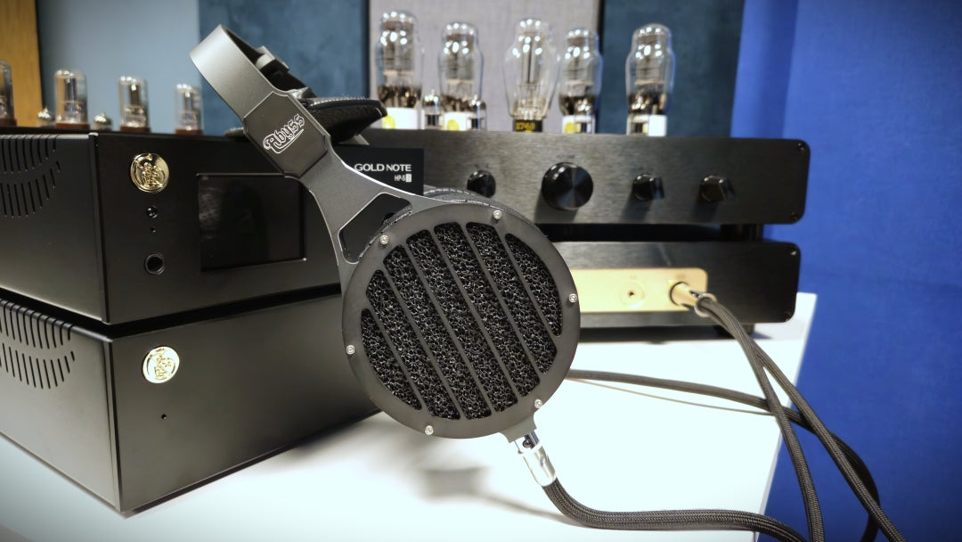 A pair of over-ear headphones with a circular mesh grille and a black headband, resting on top of black audio equipment with vacuum tubes in the background.